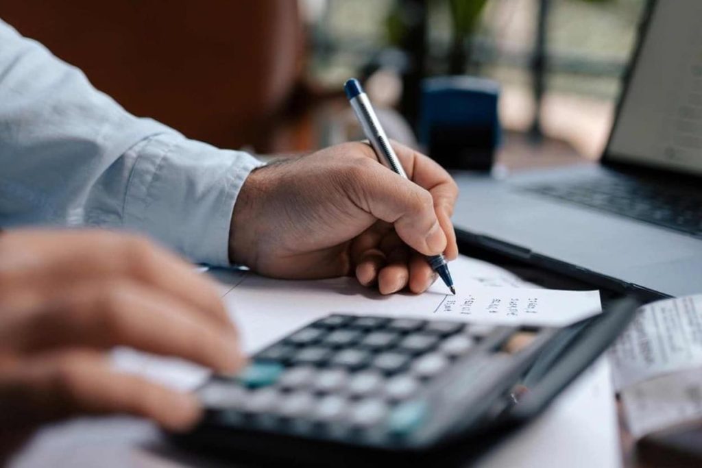 Revenue Cycle Management Collections: How Agencies Improve Medical Billing Process 3 Person writing on paper with a pen while using a calculator on a desk