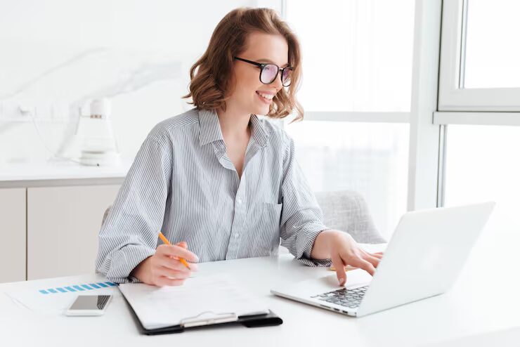 Accounts Receivable vs Accounts Payable: Key Differences 3 Smiling woman with glasses taking notes on a clipboard while working on a laptop at a bright desk.