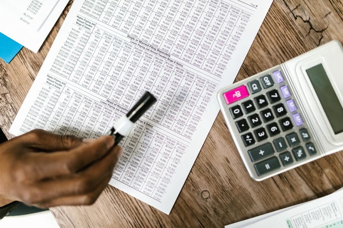 Accounts Receivable vs Accounts Payable: Key Differences 4 Hand holding a pen over printed financial tables beside a calculator on a wooden desk.